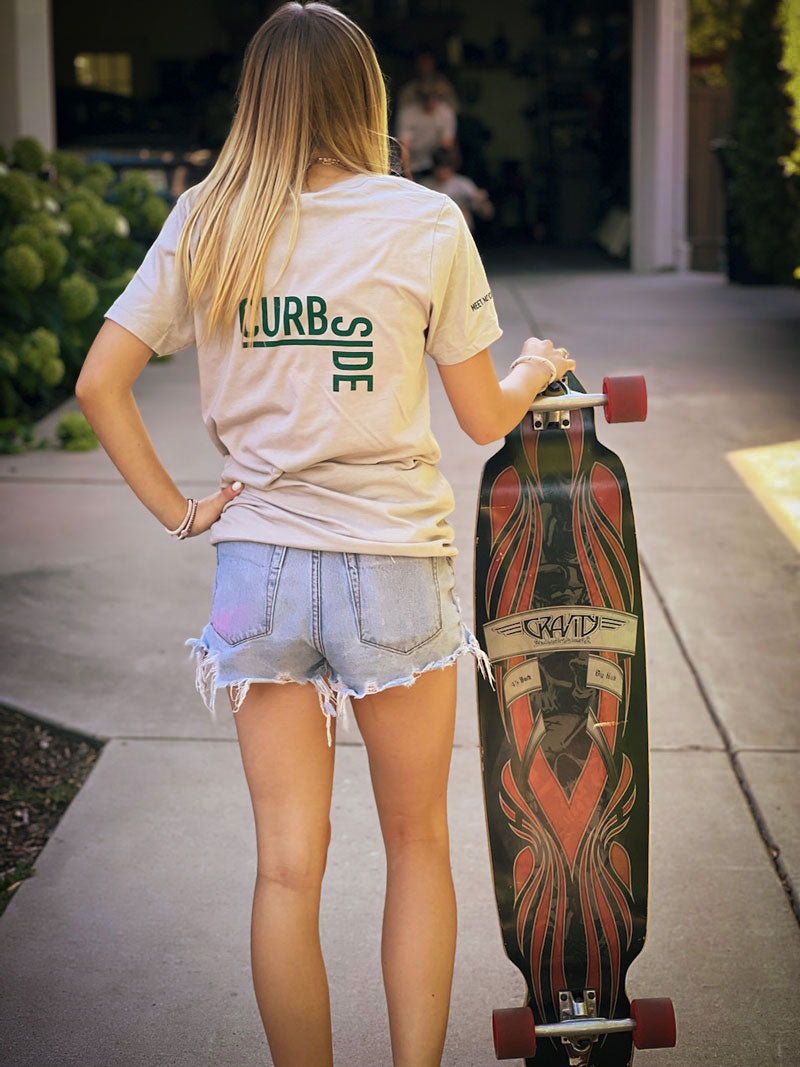 teenage girl holding a longboard skateboard with her back to camera wearing a soft t shirt with curbside logo on the back